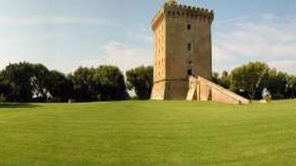 Ampio prato verde in un parco, con alberi lungo i bordi e una torre in mattoni con merlature e scalinata in pietra al centro. Sullo sfondo si vede una fila di alberi e alcuni piccoli edifici bassi sulla destra.