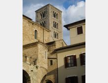 Torre campanaria in pietra di una chiesa in stile romanico, con finestre ad arco e muratura a blocchi, affiancata da un edificio residenziale chiaro con persiane in legno. In basso si vede un’arcata in pietra e, sullo sfondo, il cielo parzialmente nuvoloso.