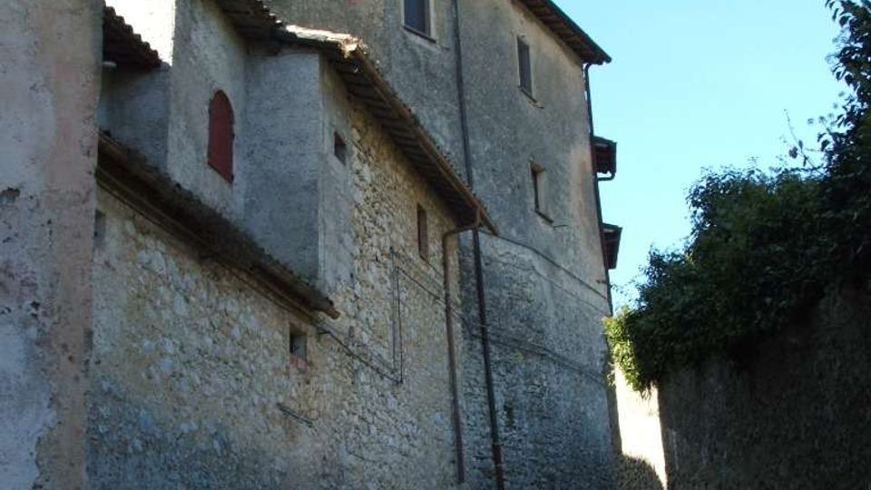 Vicolo stretto e in pendenza tra edifici in pietra e intonaco con piccole finestre e tetti a falda, illuminato da un lampione a muro. A destra corre un muretto con vegetazione, sotto un cielo sereno.