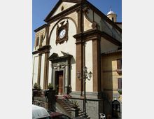 Facciata di una chiesa storica con intonaco chiaro e cornici in pietra scura, grande orologio centrale e portale rialzato con scalinata e ringhiera. Davanti si vedono un lampione a tre lanterne, vasi con piante e case adiacenti sotto un cielo sereno.