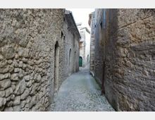 Vicolo stretto in un borgo storico, con pavimentazione in ciottoli e alte pareti in pietra grezza ai lati. In fondo si intravedono archi e porte di edifici in muratura lungo la strada curva.