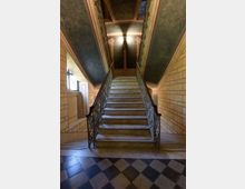 Scalinata interna in pietra che sale verso un pianerottolo, con ringhiere in ferro battuto su entrambi i lati. Pareti decorate con motivi geometrici, soffitto inclinato e pavimento a scacchiera in primo piano.