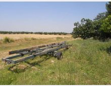 Campo rurale con erba alta e una lunga struttura metallica su ruote appoggiata a terra in primo piano. Sullo sfondo si vedono distese di campi coltivati e una linea di alberi sotto un cielo sereno.