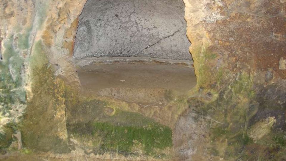 Nicchia ad arco scavata in una parete di pietra grezza, con intonaco consumato e macchie di umidità verde. La superficie è irregolare e mostra segni di deterioramento e depositi.