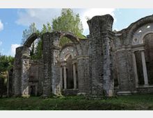 Rovine di un edificio in pietra, con grandi archi e colonne ancora in piedi e aperture ad arco con oculi circolari. L’area è circondata da vegetazione e alberi, con prato in primo piano e cielo visibile sullo sfondo.
