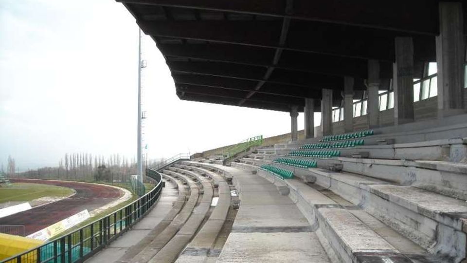 Tribuna coperta di uno stadio con gradinate in cemento e file di sedili verdi, affacciata su una pista di atletica curva. Struttura con tettoia scura sostenuta da pilastri e parapetti metallici lungo il bordo della pista.