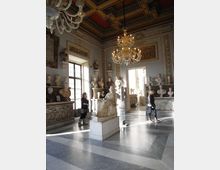 Interno di una sala museale con pavimento in marmo a motivi geometrici, pareti ornate da rilievi e busti scultorei, e una statua seduta su piedistallo al centro. Dal soffitto a cassettoni decorato pendono grandi lampadari, e ampie finestre e porte fanno entrare luce naturale.