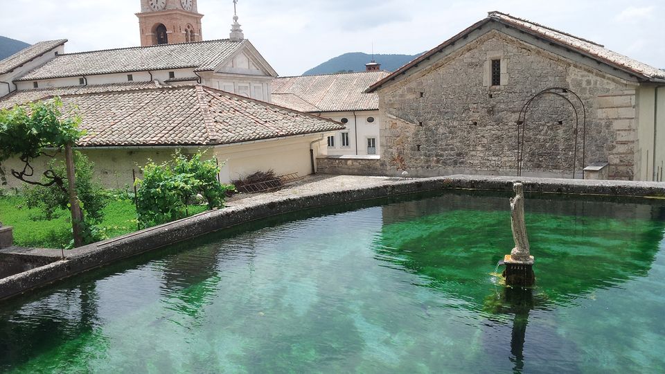 Vasca d’acqua limpida color verde smeraldo in un borgo storico, circondata da edifici in pietra e tetti in coppi. Sullo sfondo si vede un campanile con orologio e colline basse sotto un cielo nuvoloso.