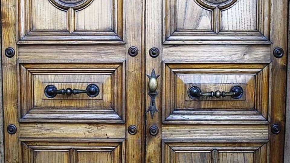 Portone in legno a doppia anta con pannelli intagliati geometrici e medaglioni circolari centrali, rifinito con borchie e maniglie in metallo scuro. Vista ravvicinata della superficie lignea con venature ben visibili e struttura simmetrica.