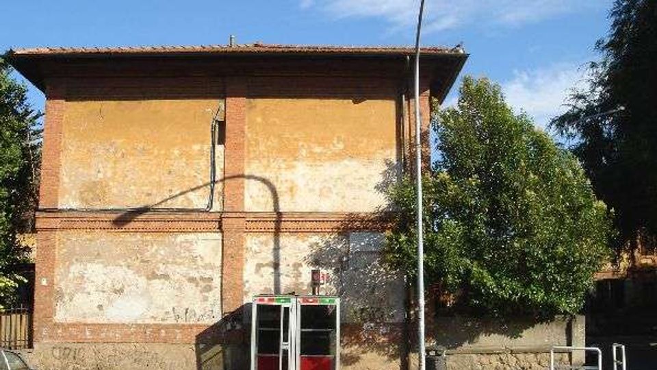 Edificio a due piani in muratura con intonaco scrostato e cornici in mattoni, affacciato su una strada. Davanti all’ingresso con piccola pensilina vetrata ci sono un lampione e alberi sul lato destro.