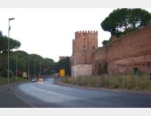 Strada asfaltata in curva con lampioni e segnaletica, affiancata da alte mura in mattoni e una torre cilindrica merlata su un basamento chiaro. Sullo sfondo si vedono alberi ad alto fusto e una zona erbosa recintata lungo il bordo della carreggiata.