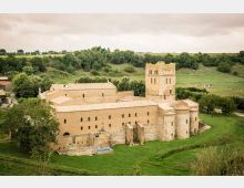 Complesso monastico in pietra con tetti in coppi e una torre campanaria quadrata, circondato da prati e alberi in una zona rurale. Sullo sfondo si vedono colline verdi e un cielo nuvoloso.
