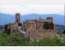 Complesso fortificato in pietra su un crinale collinare, con mura e edifici in muratura e una torre campanaria quadrata con aperture ad arco. Sullo sfondo si vedono montagne azzurrate e cielo chiaro, con vegetazione in primo piano.