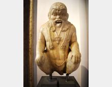 Scultura in pietra esposta su supporti in un ambiente museale, raffigurante una figura accovacciata con barba e capelli scolpiti. Sfondo con parete chiara e base espositiva scura.