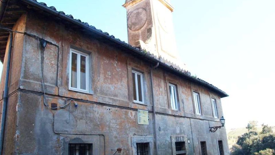 Edificio in muratura intonacata e consumata con tetto a falde e finestre rettangolari, affacciato su un panorama collinare. Dietro la copertura si innalza una torre con quadrante di orologio e piccoli elementi decorativi sulla sommità.