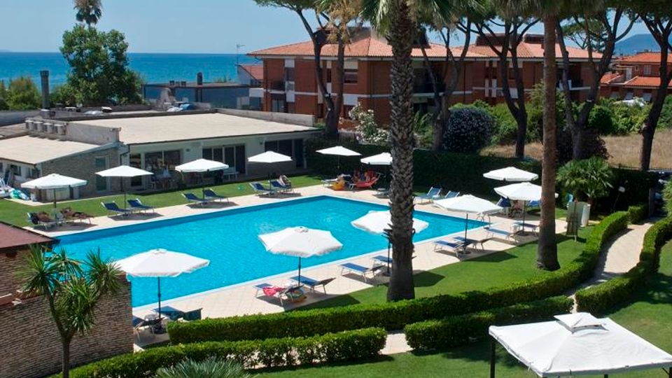 Piscina all’aperto di un hotel con lettini e ombrelloni bianchi disposti lungo il bordo, circondata da prato curato e siepi. Palme e pini marittimi incorniciano l’area, con edifici del complesso e il mare visibile sullo sfondo.