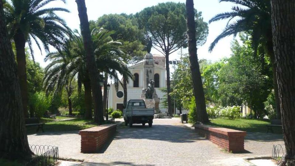 Viale pedonale in un parco, fiancheggiato da alte palme e aiuole, con muretti in mattoni lungo il percorso. Sullo sfondo si vede un edificio chiaro in stile classico con scalinata e una statua davanti.