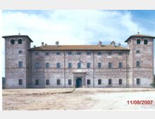 Facciata di un grande castello in mattoni con due torri quadrate laterali, tetto in coppi e file regolari di finestre. Al centro si vede un ingresso ad arco con portale, davanti a un ampio spiazzo in terra battuta sotto un cielo parzialmente nuvoloso.