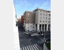 Incrocio urbano con strisce pedonali e traffico, visto dall’alto, affiancato da edifici in muratura e pietra. In primo piano un grande palazzo con portico a colonne e facciate con finestre rettangolari lungo una strada stretta fiancheggiata da palazzi.