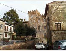 Veduta di un borgo con strada stretta e case in pietra, con auto parcheggiate ai lati. Sullo sfondo si distingue un edificio storico con facciata in mattoni e pietra, finestre ad arco, torre merlata e un piccolo orologio, circondato da alberi e cavi elettrici sospesi.