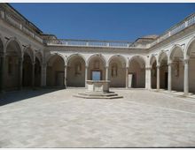 Cortile interno in pietra con ampio porticato ad archi su colonne lungo tre lati e balaustra superiore. Al centro si trova un pozzo in marmo su basamento, con nicchie e statue sulle pareti sotto un cielo sereno.