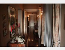 Corridoio interno di una casa con pavimento in legno scuro lucido, pareti color terra e porte bianche lungo il passaggio. A sinistra c’è un mobile con specchio, candelabri e telefono, mentre a destra tende pesanti e lampade a parete illuminano l’ambiente.