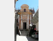 Facciata di una piccola chiesa in stile barocco alla fine di una stretta strada, con scalinata in pietra che conduce al portale e una finestra sopra l’ingresso. Impalcature e teli da cantiere circondano la parte alta dell’edificio e il lato destro della via.