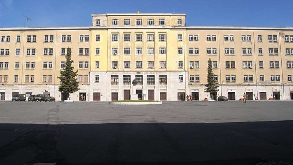 Ampia piazza asfaltata davanti a un grande edificio istituzionale rettangolare a più piani, con facciata color crema, molte finestre regolari e ingresso centrale con colonne. Ai lati dell’ingresso si vedono alcuni alberi alti e il cielo sereno sullo sfondo.