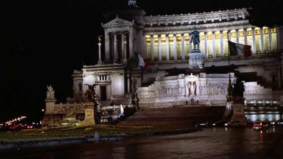 Veduta notturna del Monumento a Vittorio Emanuele II (Altare della Patria) a Roma, illuminato e con lunga scalinata e statue su basamenti in primo piano. La facciata neoclassica con colonnato e frontone si staglia contro il cielo scuro, con la strada e le luci della città ai lati.