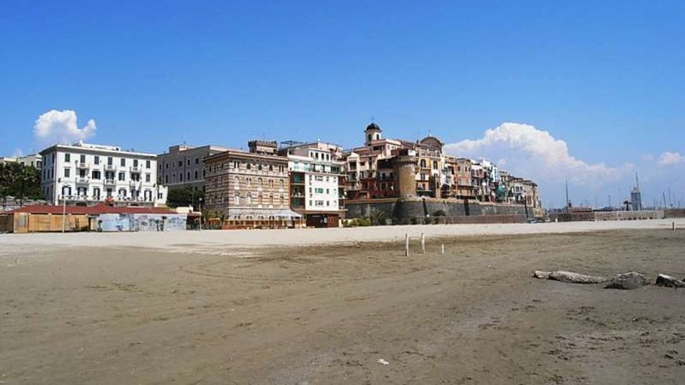 Ampia spiaggia sabbiosa in primo piano con lungomare e fila di edifici storici sullo sfondo, tra cui una torre cilindrica fortificata vicino alla costa. Cielo sereno e, sulla destra, area portuale con alberi di barche visibili in lontananza.