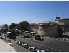Veduta di una grande fortezza costiera in pietra con bastioni curvi e bandiere sulla sommità, affacciata sul mare. In primo piano si vede una strada ampia con auto e autobus, palme lungo il marciapiede e una piccola pensilina al centro dell’incrocio.