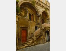 Facciata di un edificio storico in pietra con loggiato ad archi e colonne, affreschi murali consumati e vegetazione che spunta dalle cornici. Una scalinata esterna in muratura conduce al piano superiore con porte e finestre ad arco, affacciate su una strada lastricata.