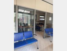 Sala d’attesa in un ufficio, con sedute blu in plastica su pavimento chiaro e pareti a vetrate con telai metallici. Dietro le vetrate si vedono stanze con scrivanie, computer, scaffali di documenti e finestre con veneziane.