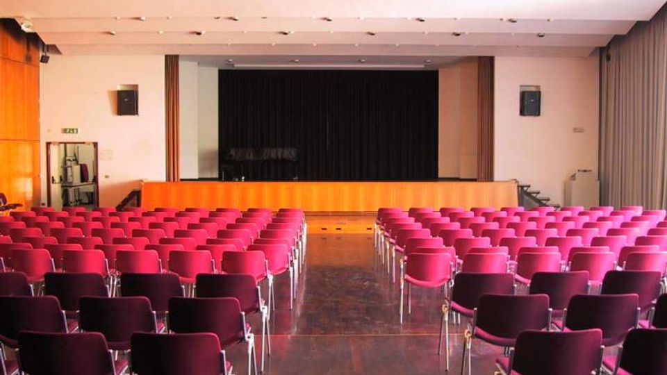 Sala auditorium con file di sedie rosse disposte a platea e corridoio centrale, rivolta verso un palco rialzato con pannellatura in legno e fondale scuro. Soffitto con illuminazione a incasso, pareti chiare e tende laterali.