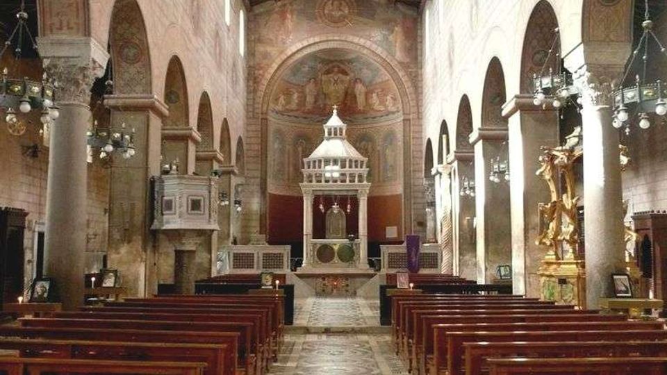 Interno di una chiesa con navata centrale fiancheggiata da arcate e colonne, soffitto a capriate in legno e file di panche in legno. Il pavimento a mosaico con motivi circolari conduce all’altare con ciborio, sullo sfondo di un’abside affrescata.