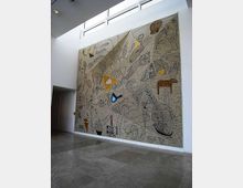 Ampio atrio interno moderno con pavimento in pietra grigia, pareti bianche e finestre alte a nastro vicino al soffitto. Una grande parete è occupata da un murale astratto su fondo beige con figure geometriche e motivi di foglie e animali stilizzati.