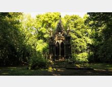Piccola cappella in pietra in stile gotico, con frontone a punta, pinnacoli e grande finestra ad arco con grata, situata in una radura. Intorno si vede un fitto bosco di alberi e vegetazione che circonda l’edificio e l’area erbosa.