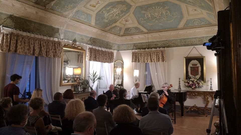 Sala storica adibita a concerto, con soffitto a volta decorato da affreschi e riquadri azzurri, pareti bianche con grandi finestre drappeggiate e specchi dorati. In fondo, un’area con pianoforte e arredi classici, tra cui una consolle intagliata con stemma incorniciato e composizioni floreali.