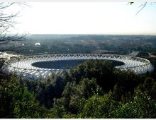 Veduta panoramica di un grande stadio ovale con copertura bianca a struttura reticolare, visto dall’alto tra alberi e vegetazione in primo piano. Sullo sfondo si estende la città con edifici e strade sotto un cielo luminoso.