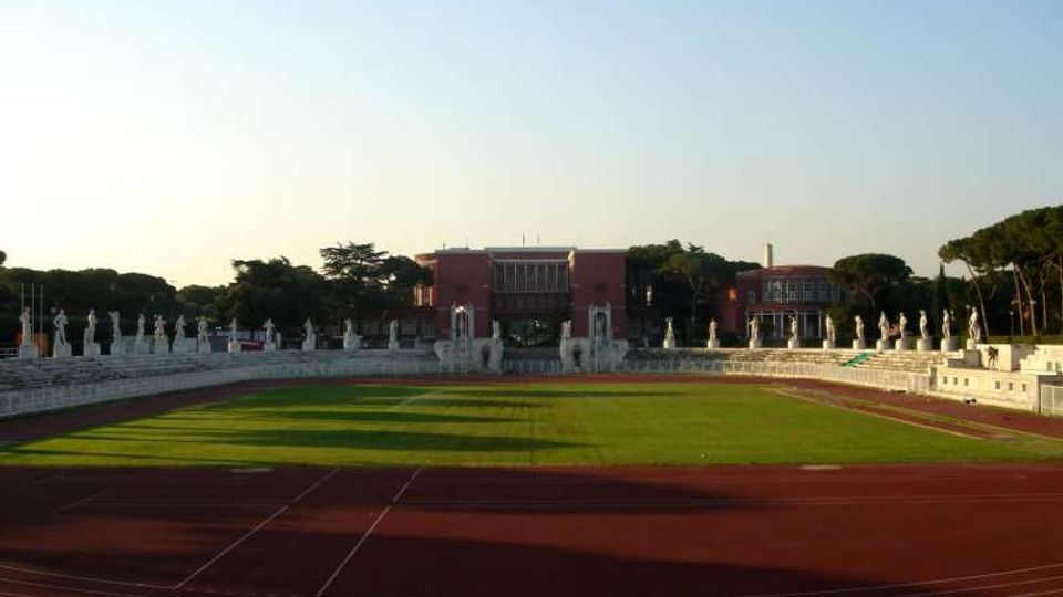 Vista frontale di uno stadio con pista di atletica rossa che circonda un campo in erba verde. Sullo sfondo si vedono gradinate in pietra con statue allineate e un edificio principale in mattoni rossi, circondato da alberi.