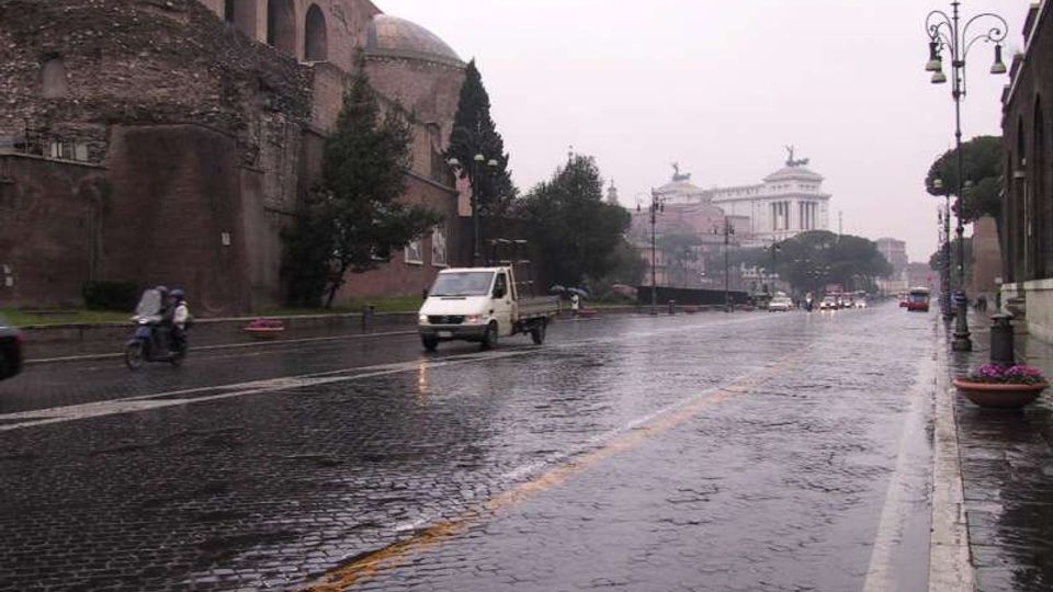 Strada urbana bagnata dalla pioggia con pavimentazione in sampietrini e lampioni lungo il marciapiede. A sinistra si vedono grandi mura in mattoni con arcate e una cupola, mentre sullo sfondo appare un edificio monumentale su una collina.