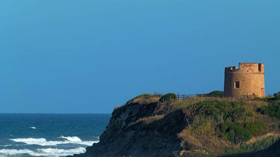 Torre costiera in pietra su un promontorio erboso affacciato sul mare, con scogliera e onde che si infrangono sulla riva sottostante. Il cielo sereno e l’orizzonte marino incorniciano la struttura isolata.