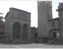 Piazza lastricata con complesso in pietra medievale, con un edificio a due arcate e scalinate d’accesso. Accanto si innalza una torre quadrata in muratura e sono visibili muri di cinta e facciate con piccole finestre.