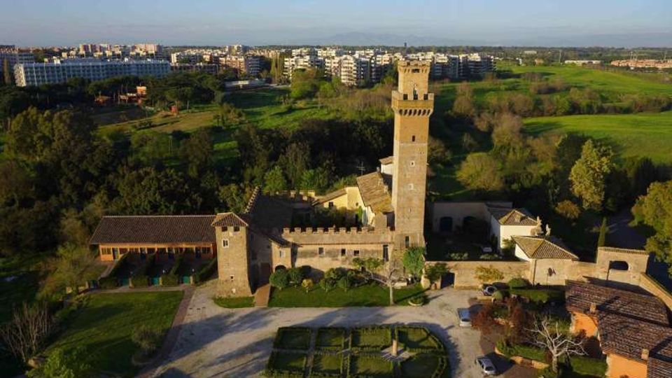 Veduta aerea di un complesso storico con torre in mattoni e mura merlate, circondato da un giardino geometrico e vialetti. Sullo sfondo si estendono campi verdi e un’area urbana con palazzi residenziali.