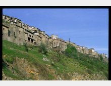 Case in pietra di un borgo arroccato lungo un pendio erboso, con edifici addossati tra loro e muri in muratura a vista. In primo piano si vede il versante roccioso con cespugli e alberi sparsi sotto un cielo sereno.