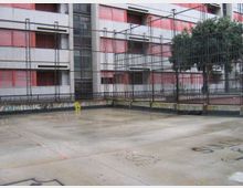 Cortile in cemento con scritte e graffiti a terra, circondato da alte recinzioni metalliche. Sullo sfondo si vede un grande edificio residenziale a più piani con balconi e persiane rosse, e un albero vicino alla rete.