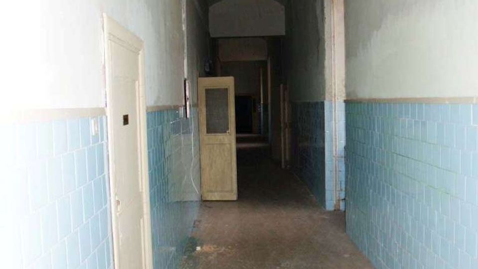 Corridoio interno lungo e stretto di un edificio, con pareti rivestite a metà da piastrelle azzurre e parte superiore intonacata chiara, pavimento in cemento e soffitto a volta. Diverse porte lungo i lati e una porta vetrata aperta in fondo al passaggio.