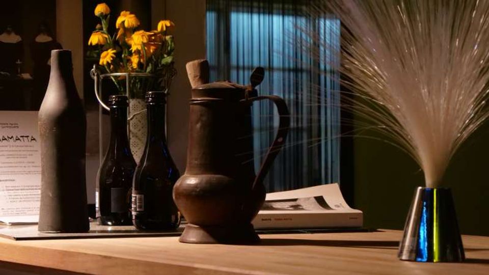 Tavolo in legno con bottiglie e un recipiente in ceramica, un vaso con fiori gialli e un libro appoggiato. Sul lato destro c’è una piccola base con filamenti decorativi, con una finestra a tendine sullo sfondo.