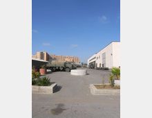 Ampio piazzale asfaltato di un’area industriale, con diversi camion parcheggiati e un grande capannone bianco con portoni sul lato destro. In primo piano ci sono aiuole con piante e una struttura circolare bassa al centro del piazzale, con edifici più alti sullo sfondo sotto un cielo sereno.