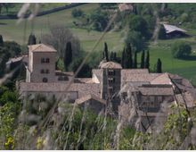 Complesso di edifici in pietra con tetti in coppi e due torri quadrate, immerso in una campagna collinare con campi verdi e filari di cipressi. In primo piano si vedono erbe e rami sfocati che incorniciano la vista dall’alto.